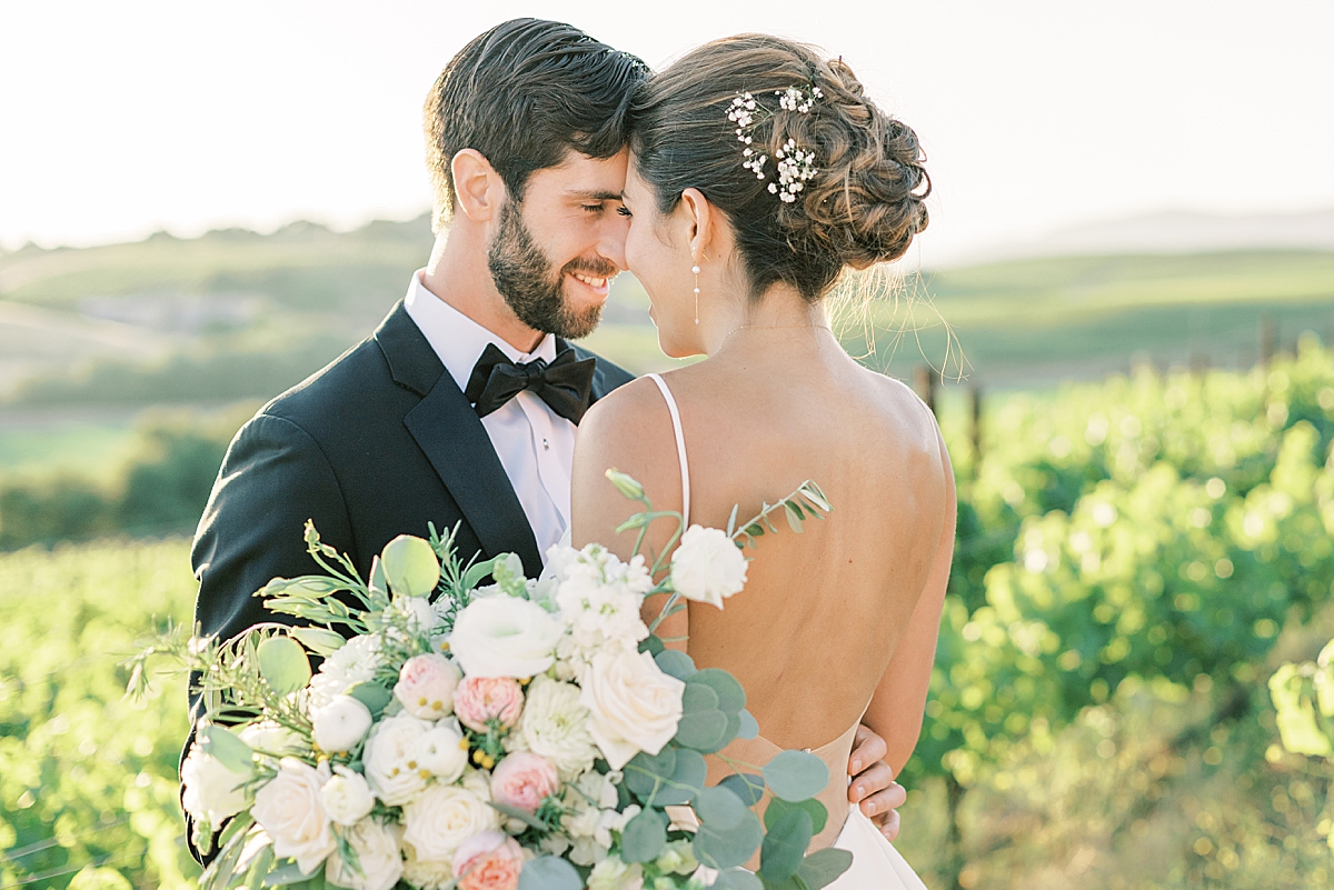 A bride and groom forehead to forehead near rows of vineyards.