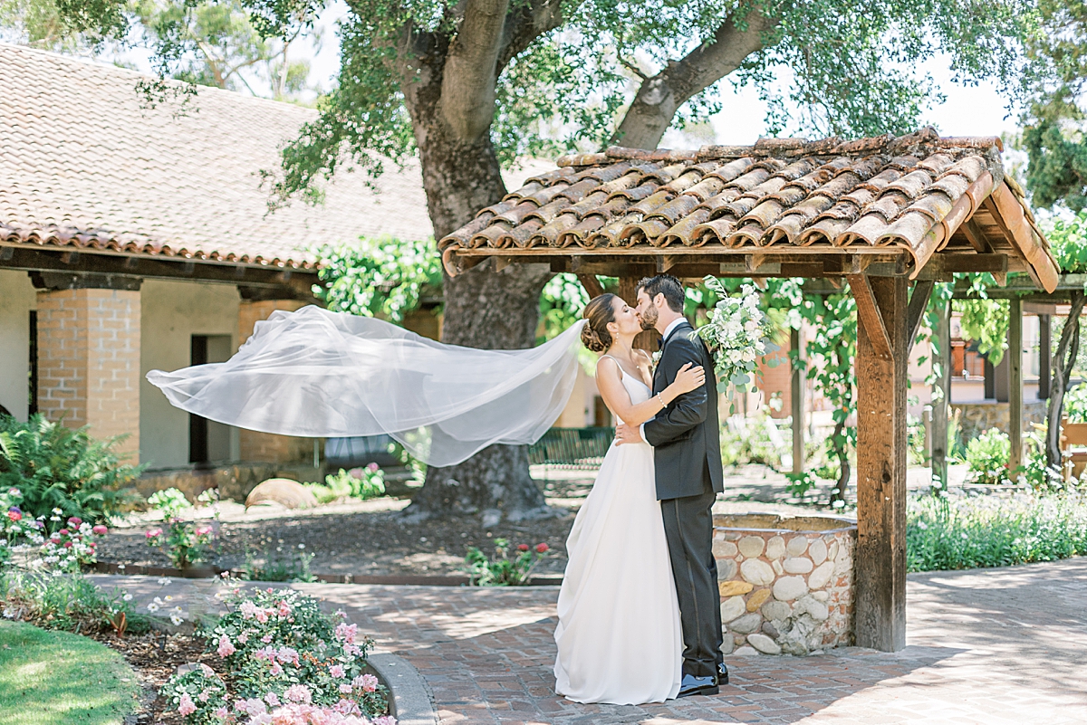 The couple sharing a kiss on the ground of the Mission in San Luis Obispo, California as the bride's veil flies in the wind behind her.