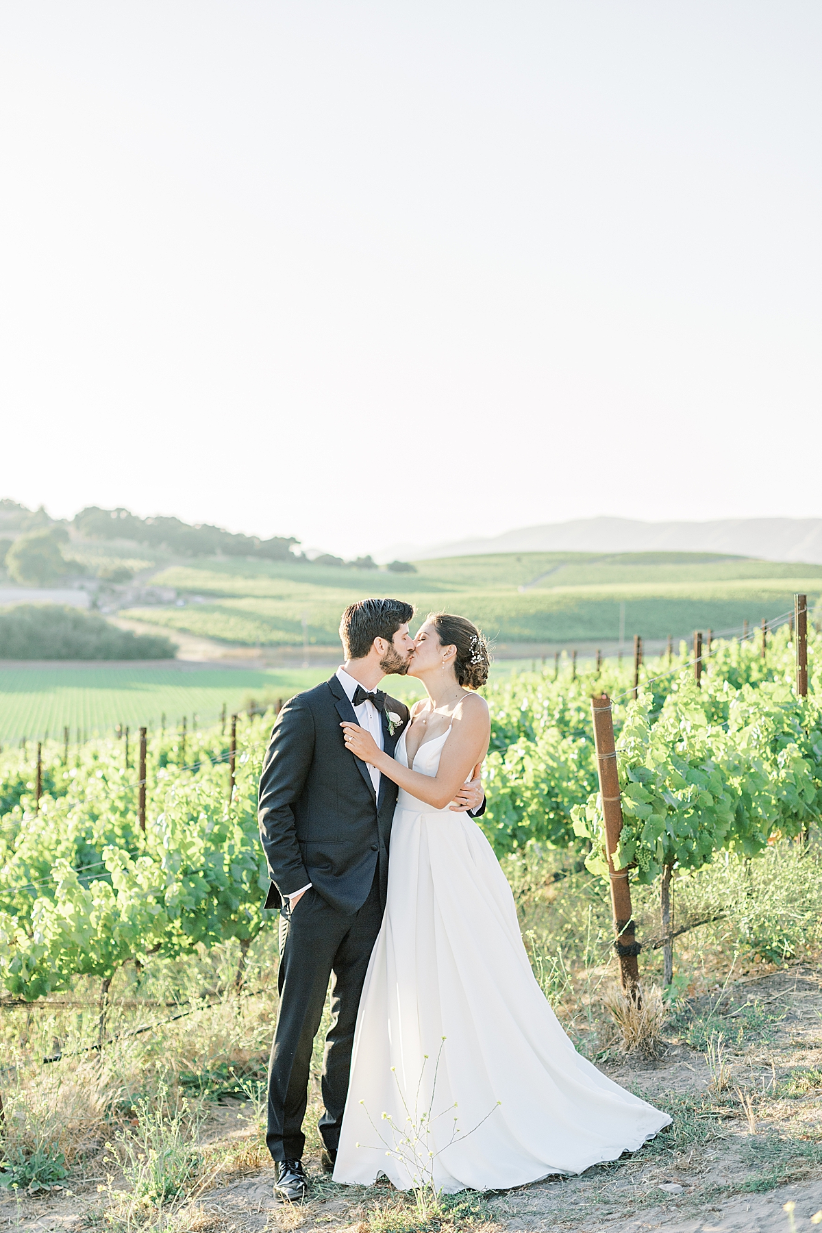 The newlyweds sharing a kiss in front of the rolling hills and vineyards during their San Luis Obispo Mission wedding day.