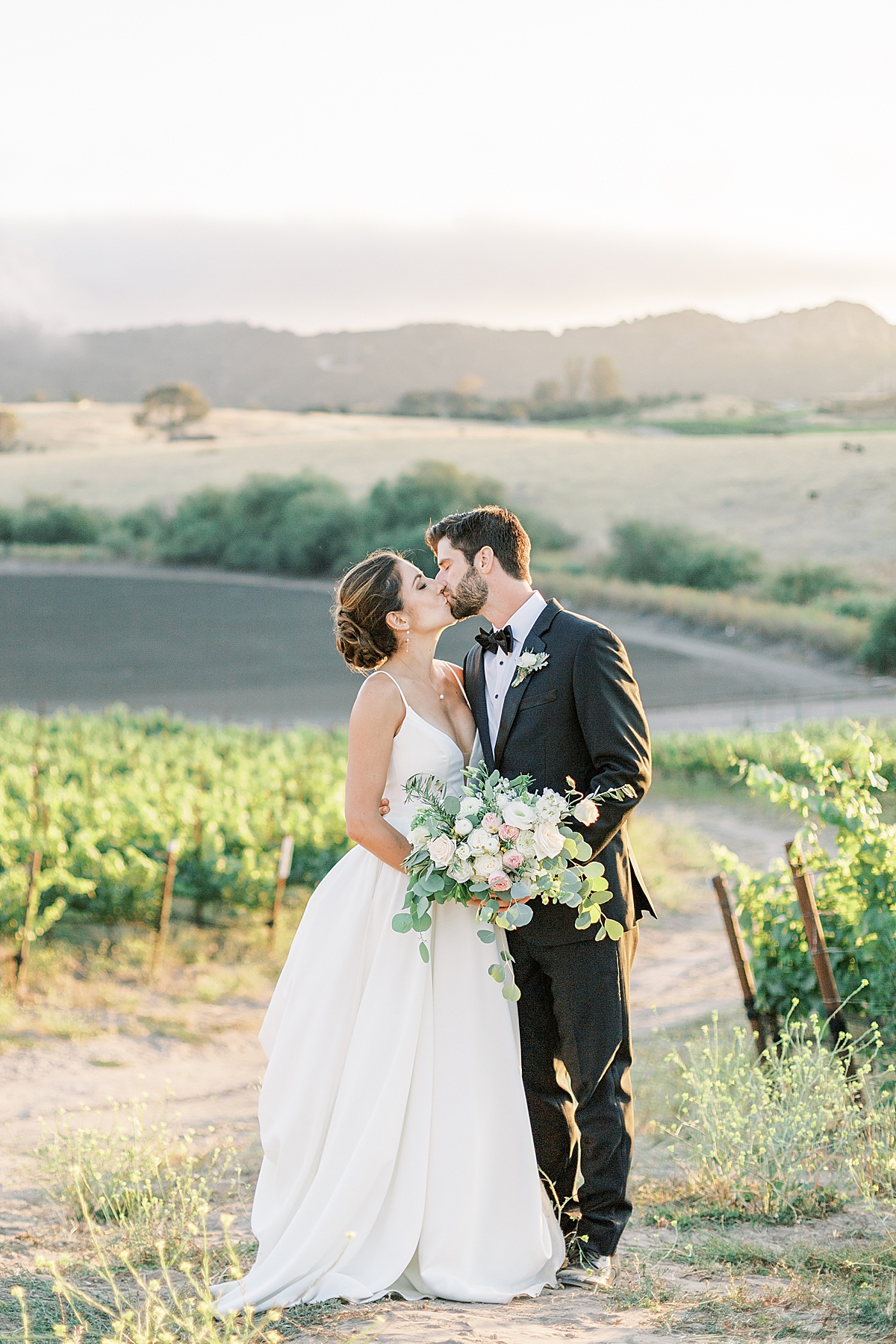 The couple sharing a kiss in front of the vineyards