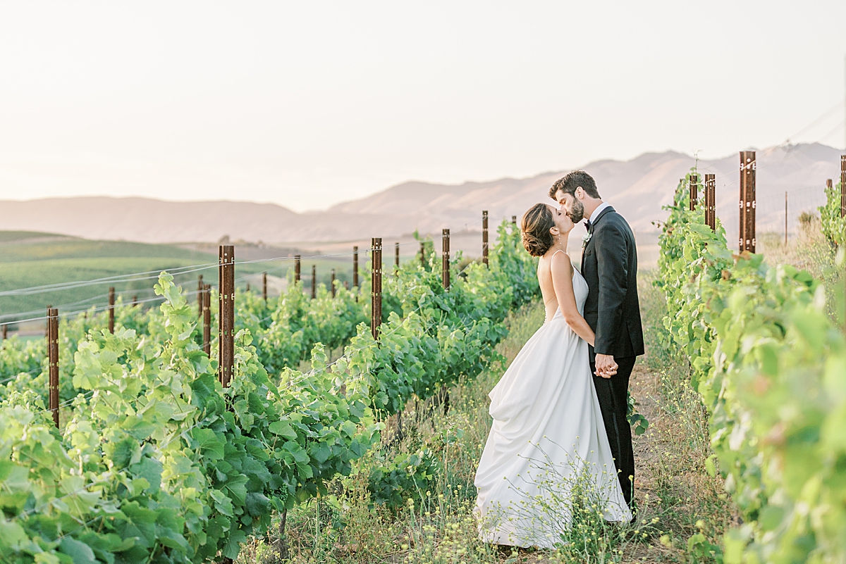The bride and groom sharing a kiss in between the rows of vines with the mountains in the background as far as the eye can see at their during their San Luis Obispo Mission wedding venue.