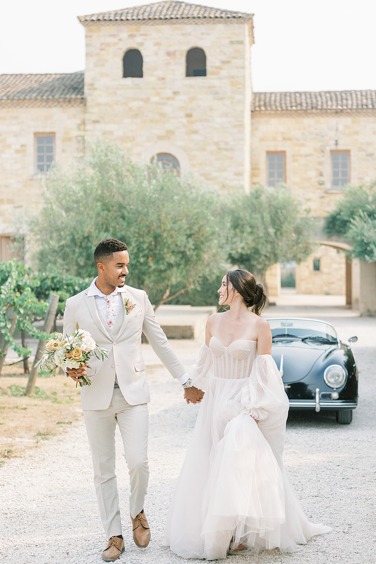 The bride and groom smiling at each other as they walk down the driveway of the Sunstone Villa. There is a vintage car behind them.