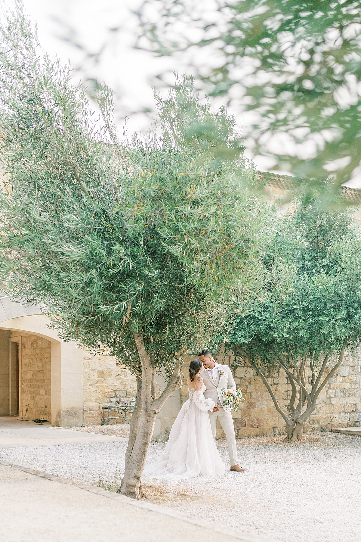 Miya & Luke sharing a kiss as he holds her bouquet under two olive trees.