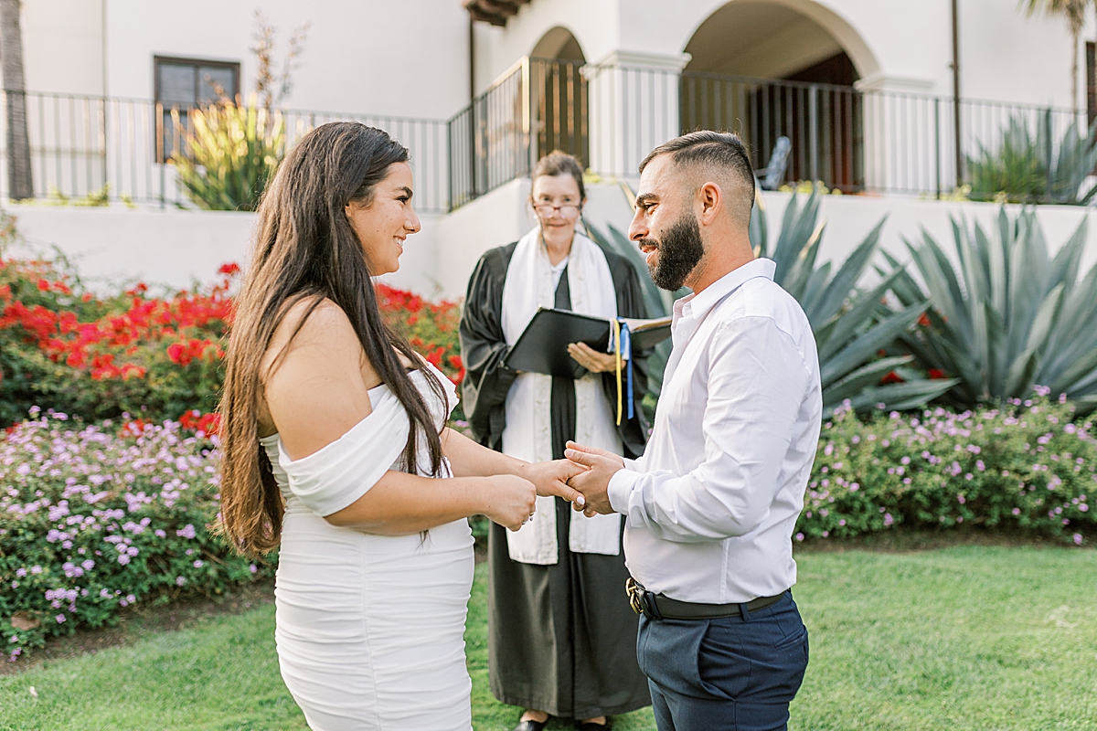 Omid placing a ring on Bella's finger as the officiant smiles in the background. 