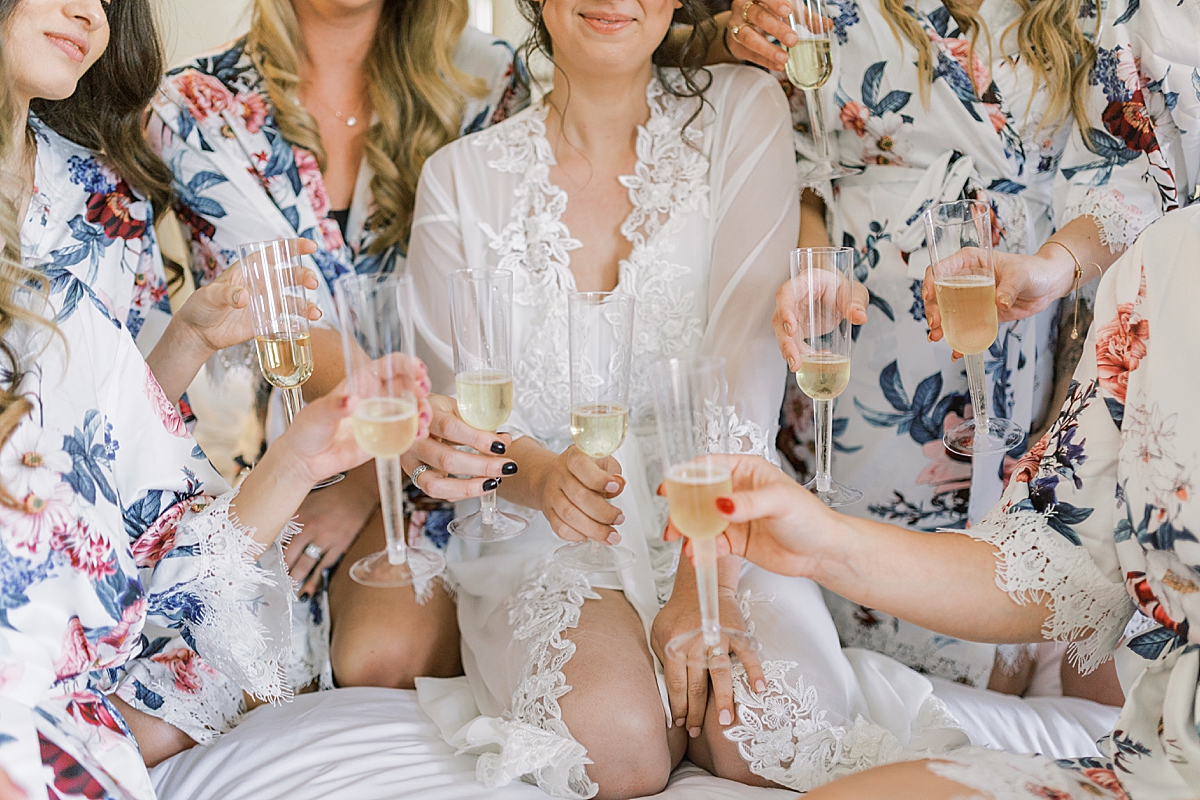 Glasses of champagne being held by the bride and her bridesmaids while wearing their matching robes and sitting on a bed.
