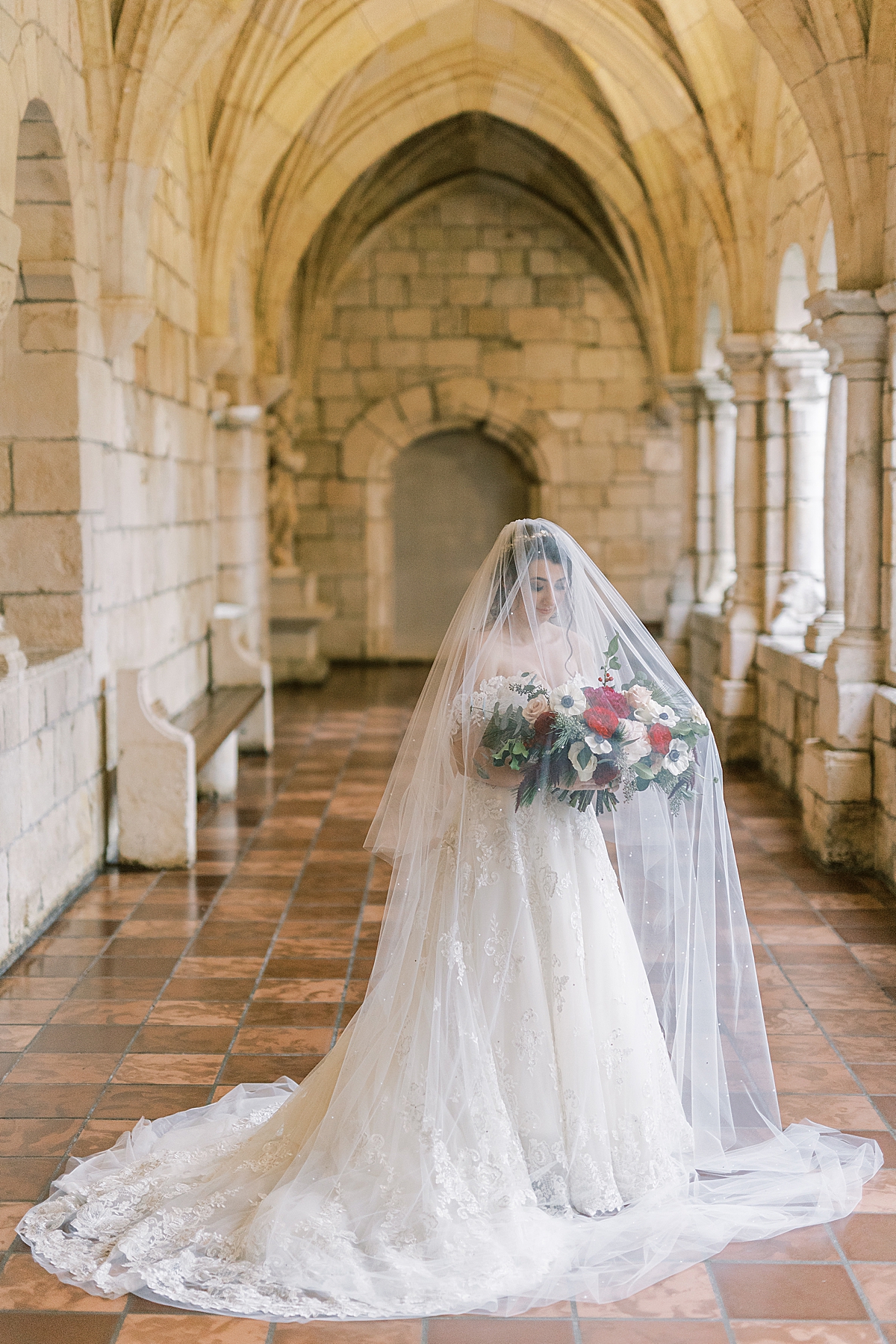 The bride standing in the halls at her Ancient Spanish Monastery Wedding venue holding her large bouquet of white and red flowers.