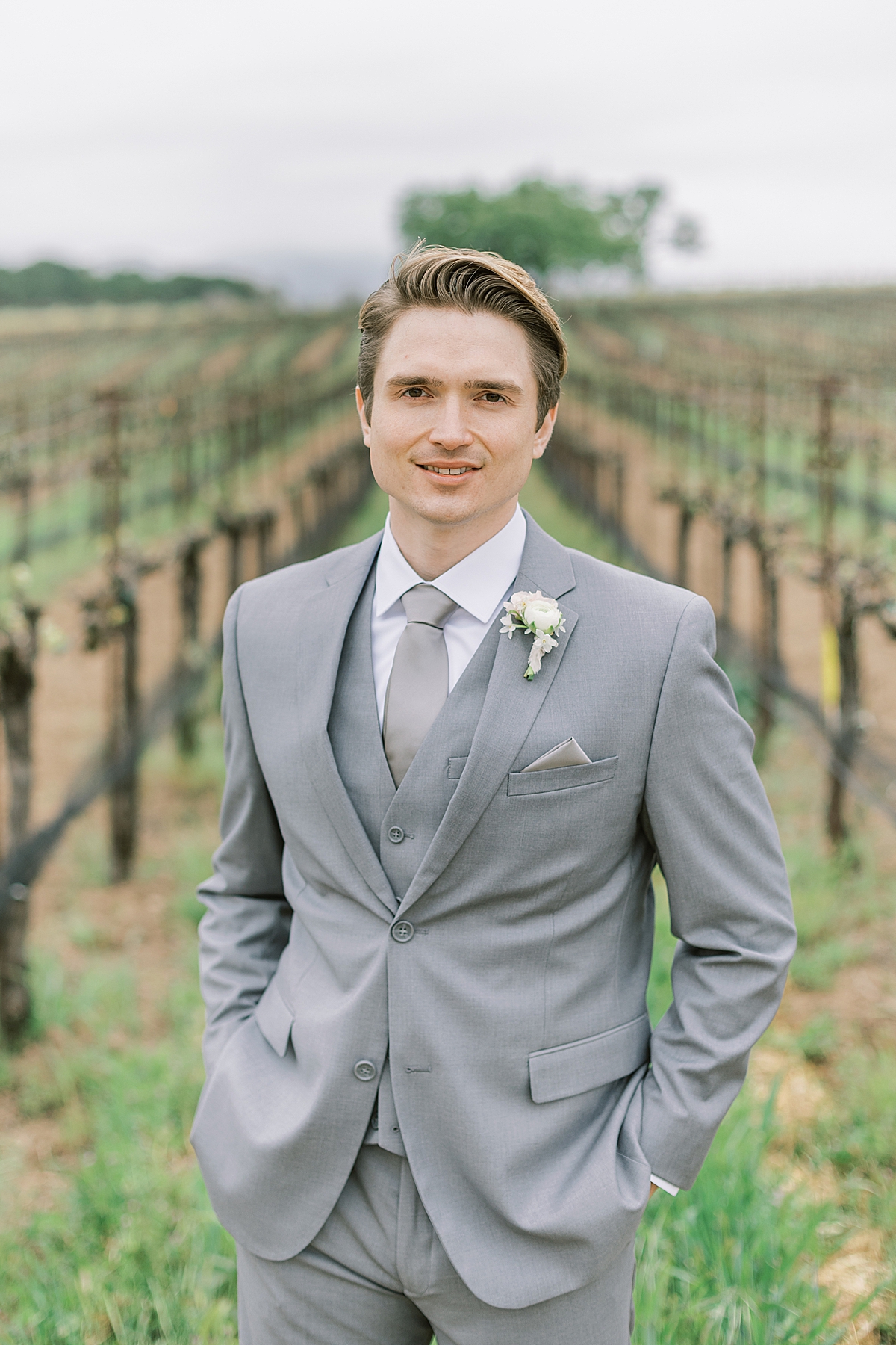 The groom smiling at the camera with rows of vineyards behind him.